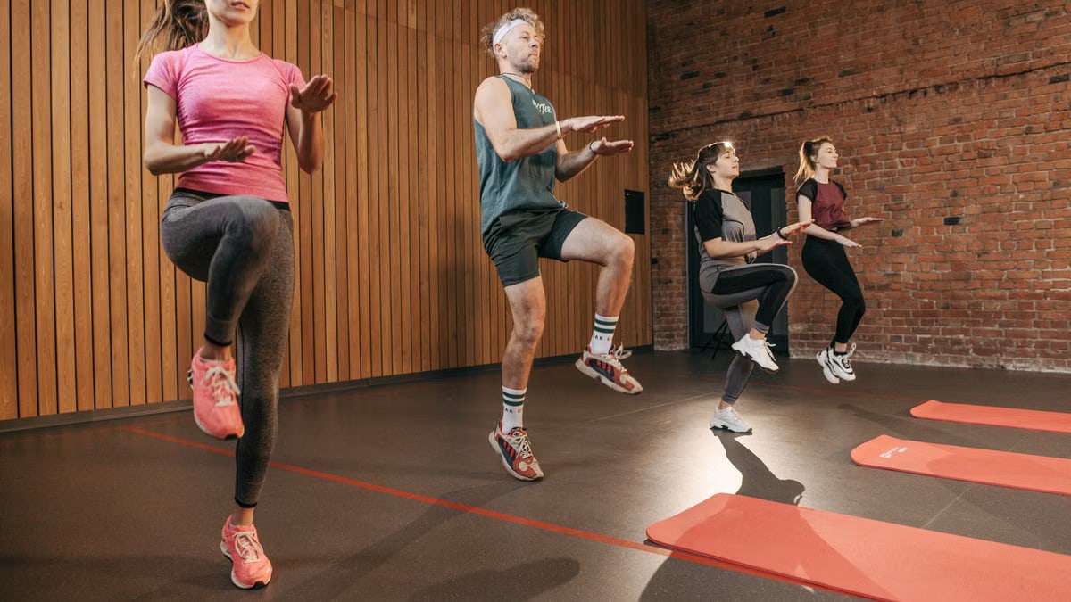 Four adults performing high knees in an indoor gym with wooden panel walls.